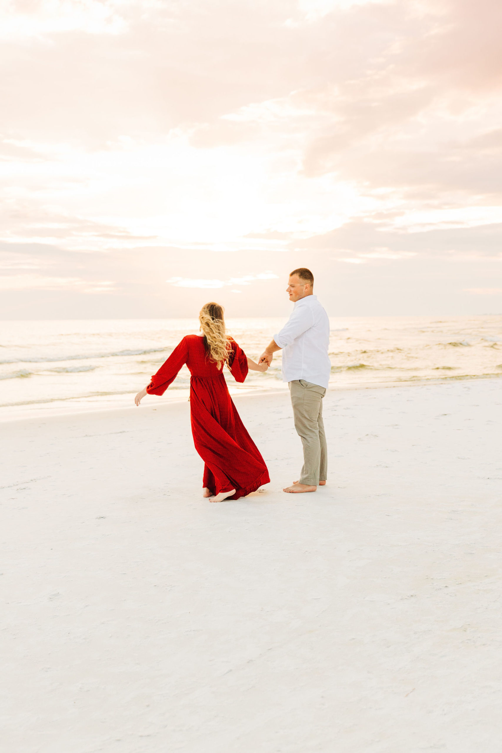 Kallie and Noah holds hands as they dance on the beach against a pink sunset